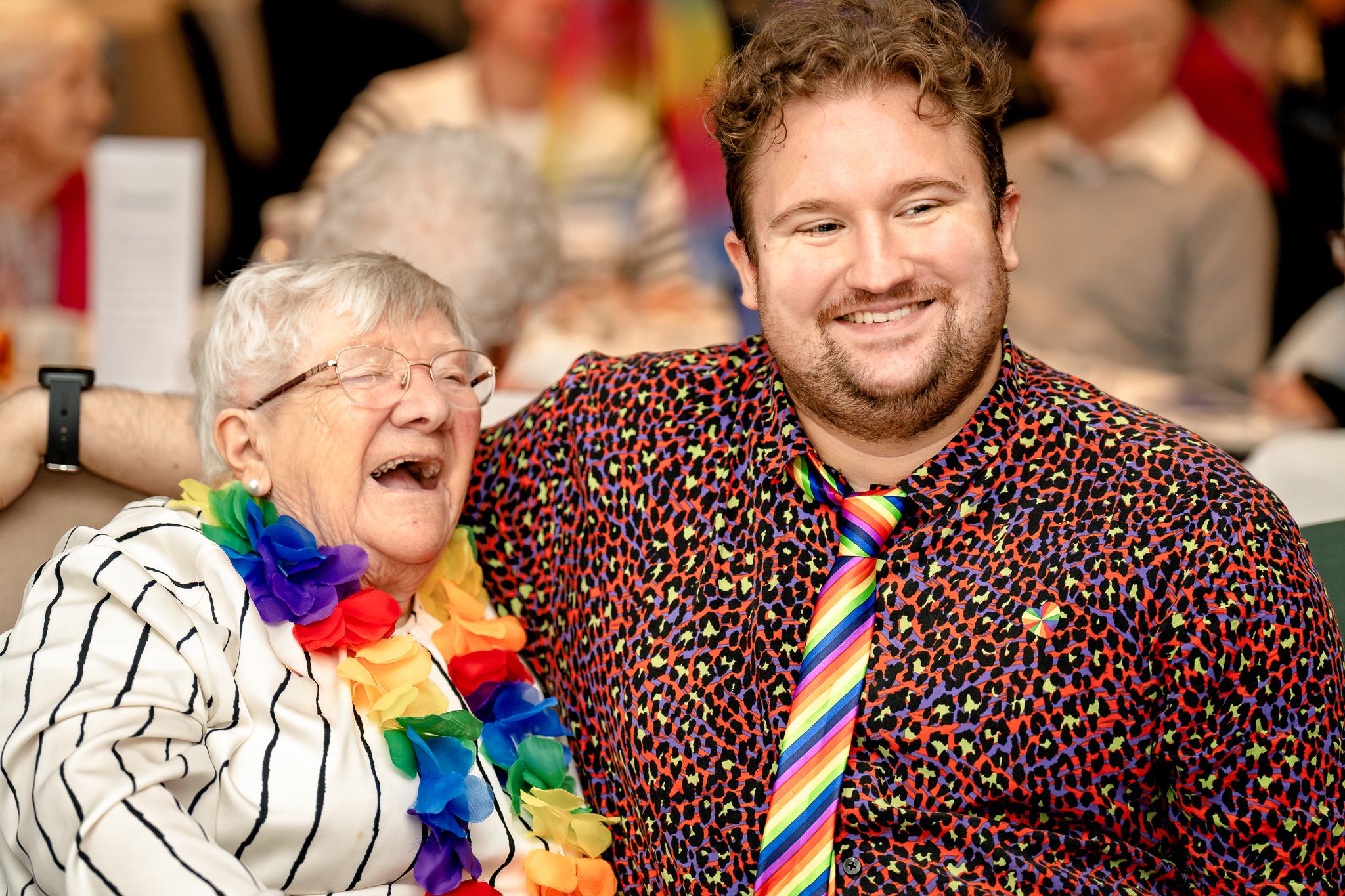 Elderly woman and man laughing together, wearing rainbow coloured accessories